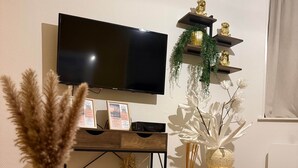Uma sala de estar moderna com uma televisão de tela plana na parede, uma mesa de centro de vidro com objetos decorativos e uma mesa lateral de madeira com uma pequena planta e uma tigela decorativa.