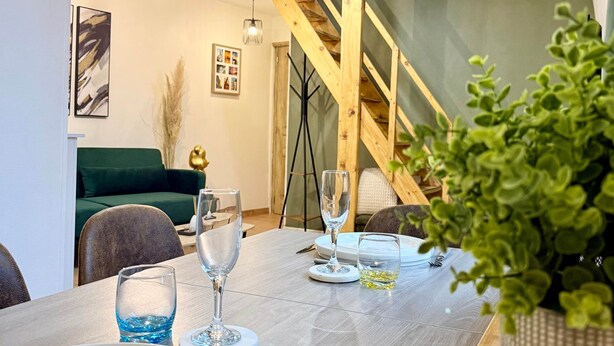 Un coin repas avec une table en bois dressée pour deux personnes, un escalier en arrière-plan et une plante verte sur la table.