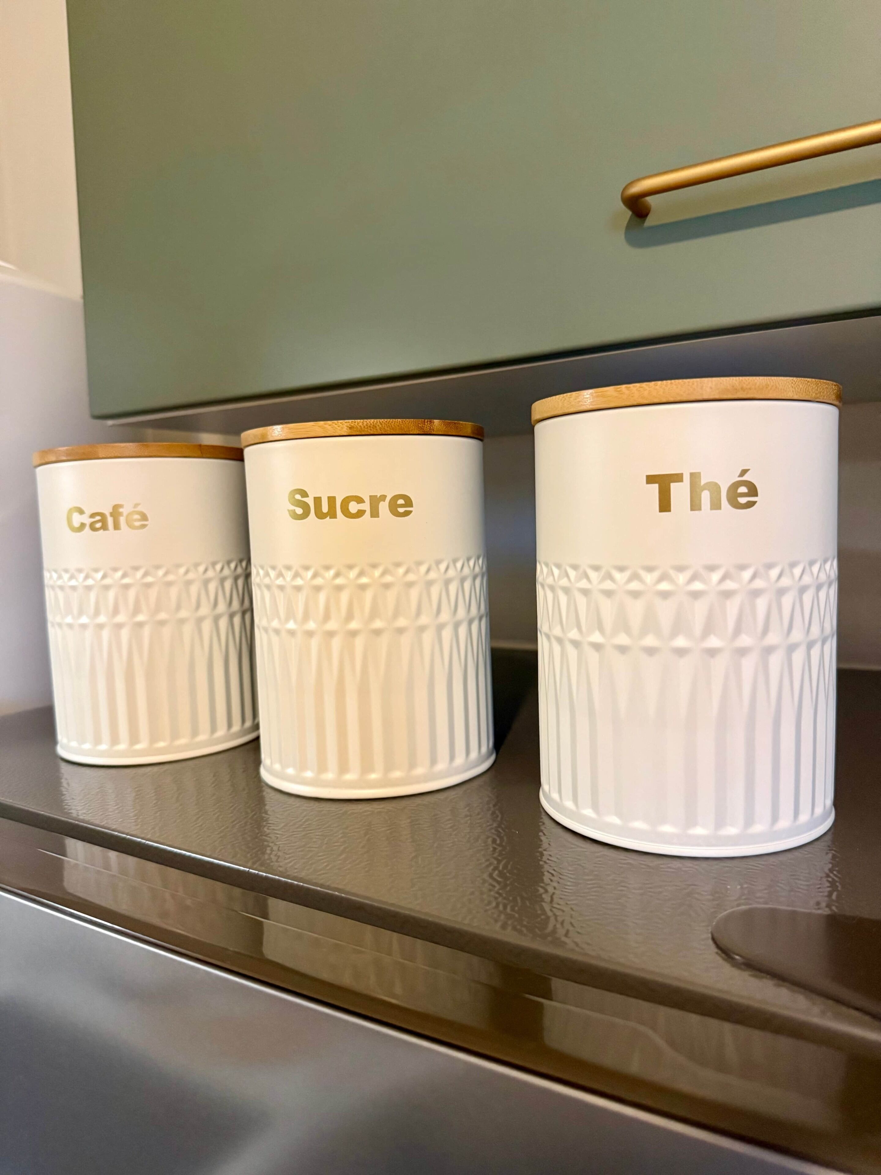 Three white containers with gold trim labeled "Café," "Sucre," and "Thé" on a shelf.
