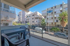 A balcony with wicker furniture, a view of a street with a white van and a car, and multi-story buildings with balconies and satellite dishes. - Gurcuoglu Homes (Antalya)
