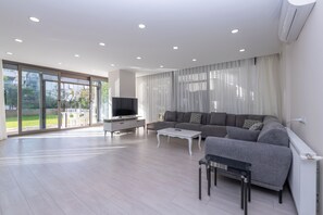 A spacious living room with a grey sofa, a white coffee table, and a TV on a stand. There is a sliding glass door leading to an outdoor area. - Gurcuoglu Homes (Antalya)