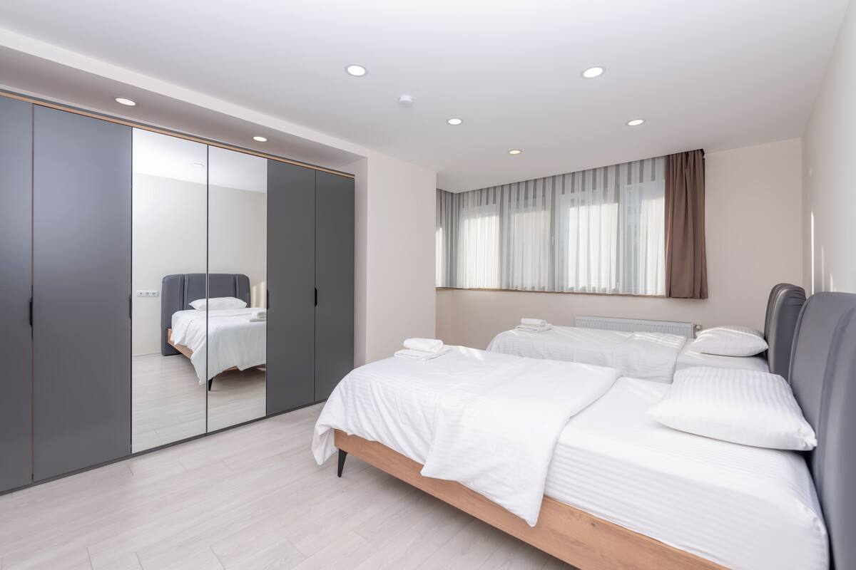 Une chambre moderne avec un grand miroir, un lit recouvert de draps blancs, une armoire grise et une fenêtre avec des rideaux.