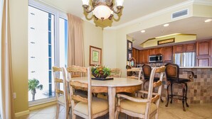 A dining area with a round table and chairs, a kitchen with wooden cabinets, and a large window with curtains. - Beachfront 4BR Silver Beach Towers West 306 (Destin)