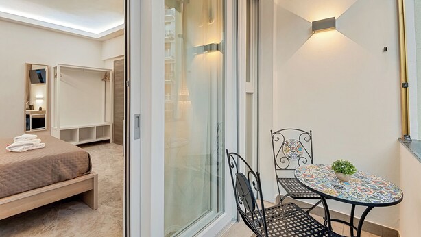 Une chambre moderne avec un lit, une armoire, une porte-fenêtre coulissante et un petit balcon équipé d’une table et de chaises.