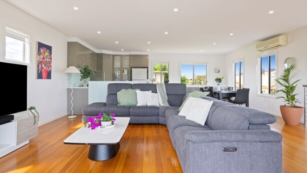 A modern living room with a grey sectional sofa, a flat-screen TV, a coffee table with flowers, and a dining area with a table and chairs. - Baravi Breeze Retreat (Safety Beach)