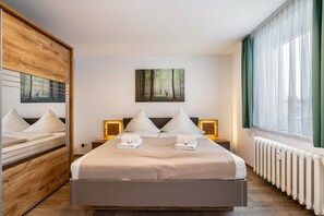 Habitación de hotel con una cama grande, un espejo, un cuadro, una ventana con cortinas y una mesita de noche con lámpara.