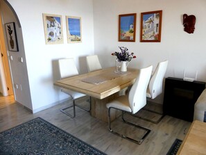 Un comedor con una mesa de madera, sillas blancas y un jarrón con flores. Hay dos cuadros en la pared.