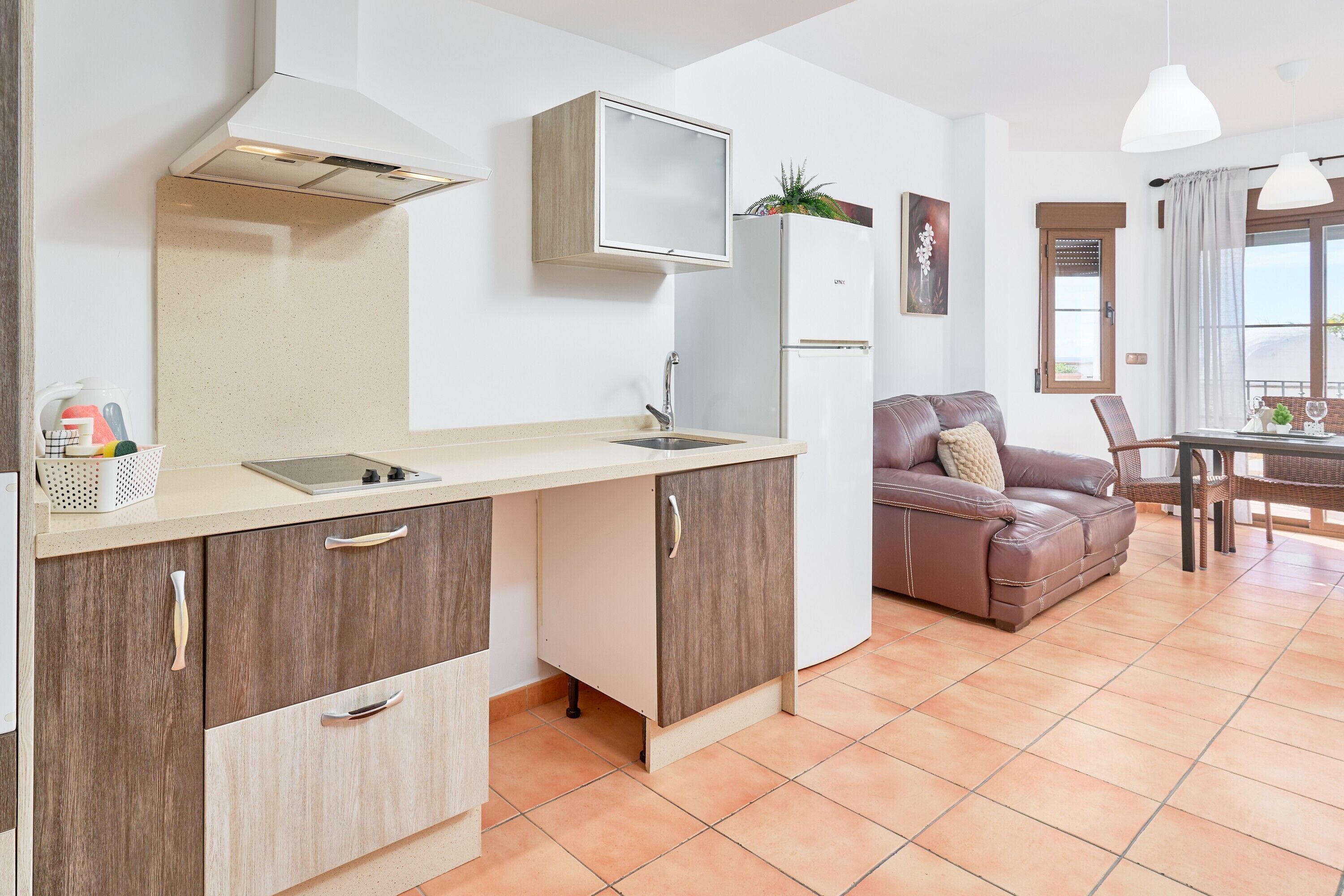 Una kitchenette con un lavamanos, armarios y un refrigerador. Hay un sofá de cuero marrón y una mesa de comedor con sillas.