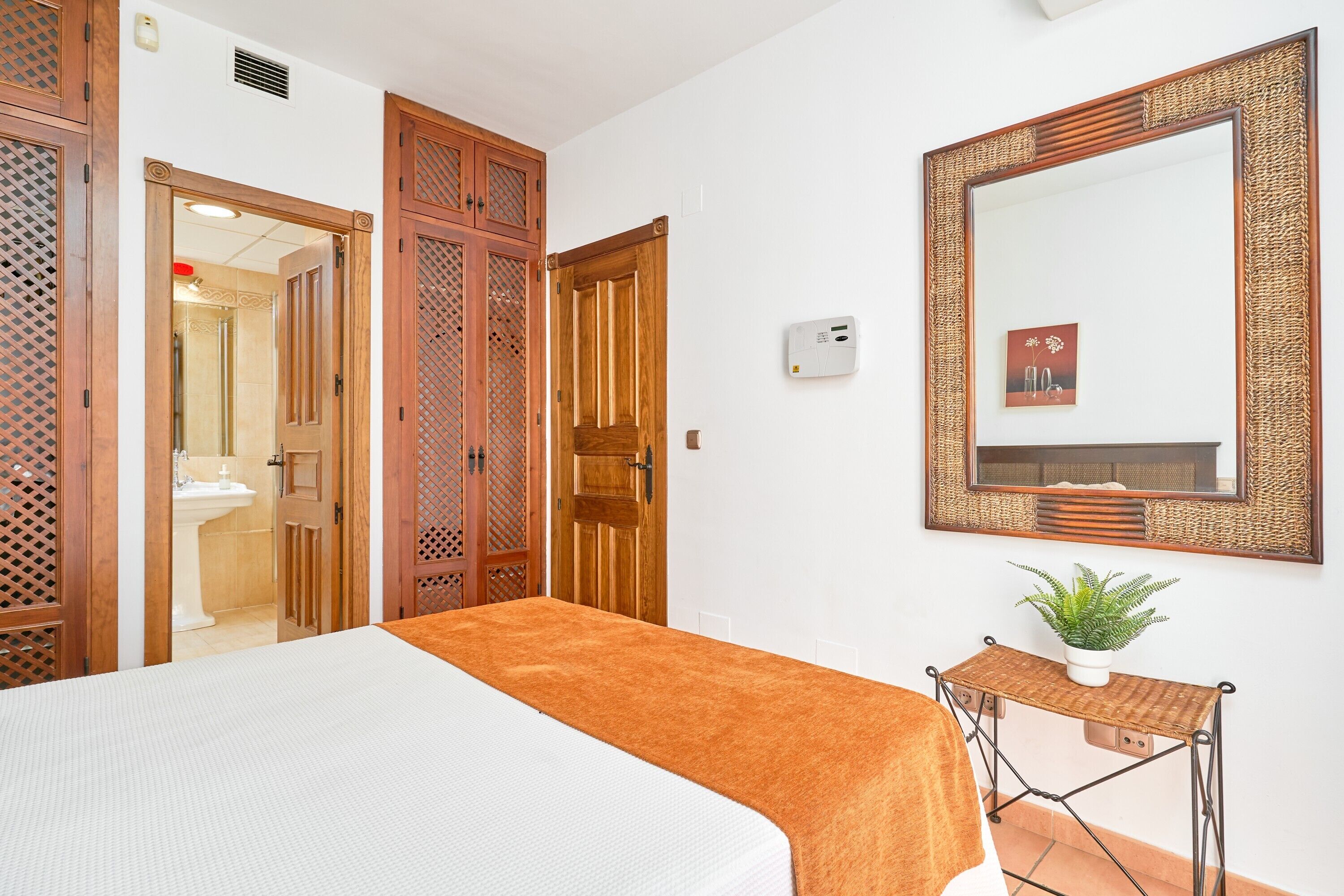 Habitación de hotel con una cama, una mesita con una planta, un espejo y puertas de madera.