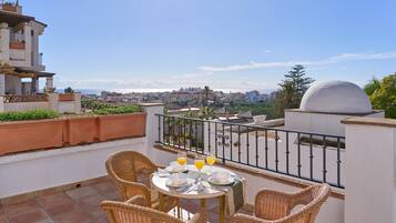Una terraza con sillas de mimbre, una mesa con un vaso de jugo de naranja y vista a un paisaje urbano.