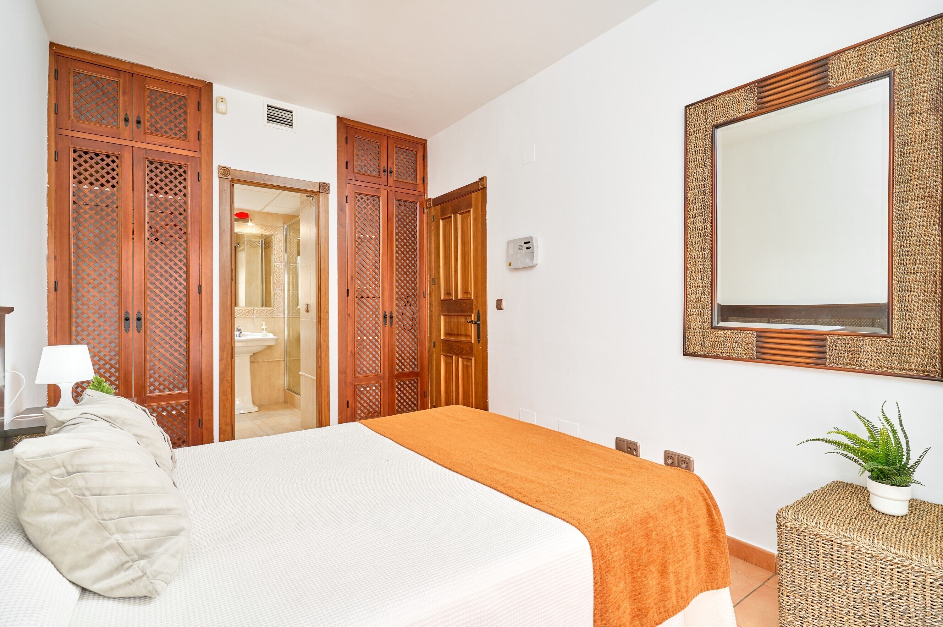 A bedroom with a bed, a bedside table, a lamp, a potted plant, and a wardrobe.