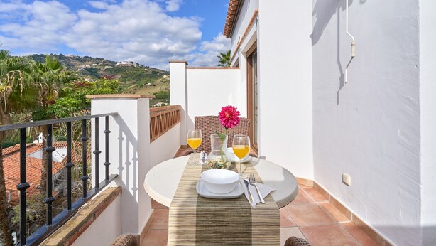 A balcony with a table set for two, a flower vase, and two glasses of orange juice.
