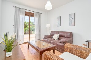 Una sala de estar con un sofá de cuero marrón, una mesa de centro de vidrio, un sillón de mimbre, una planta en maceta y un ventanal corredero que da a un balcón.
