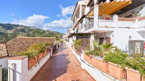 A residential building with terracotta-tiled roofs, a balcony with potted plants, and a view of a hilly landscape.