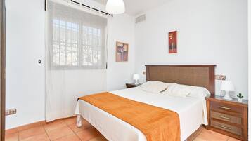 Un dormitorio con cama, mesita de noche, lámpara, una planta pequeña y ventana con cortina.