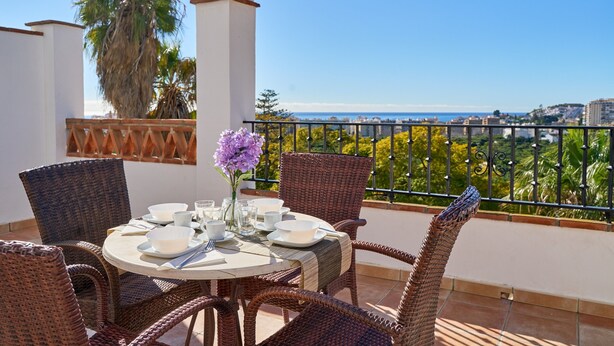 Ein runder Tisch mit Stühlen und einem Blumenstrauß auf einem Balkon mit Blick auf das Meer und Palmen.