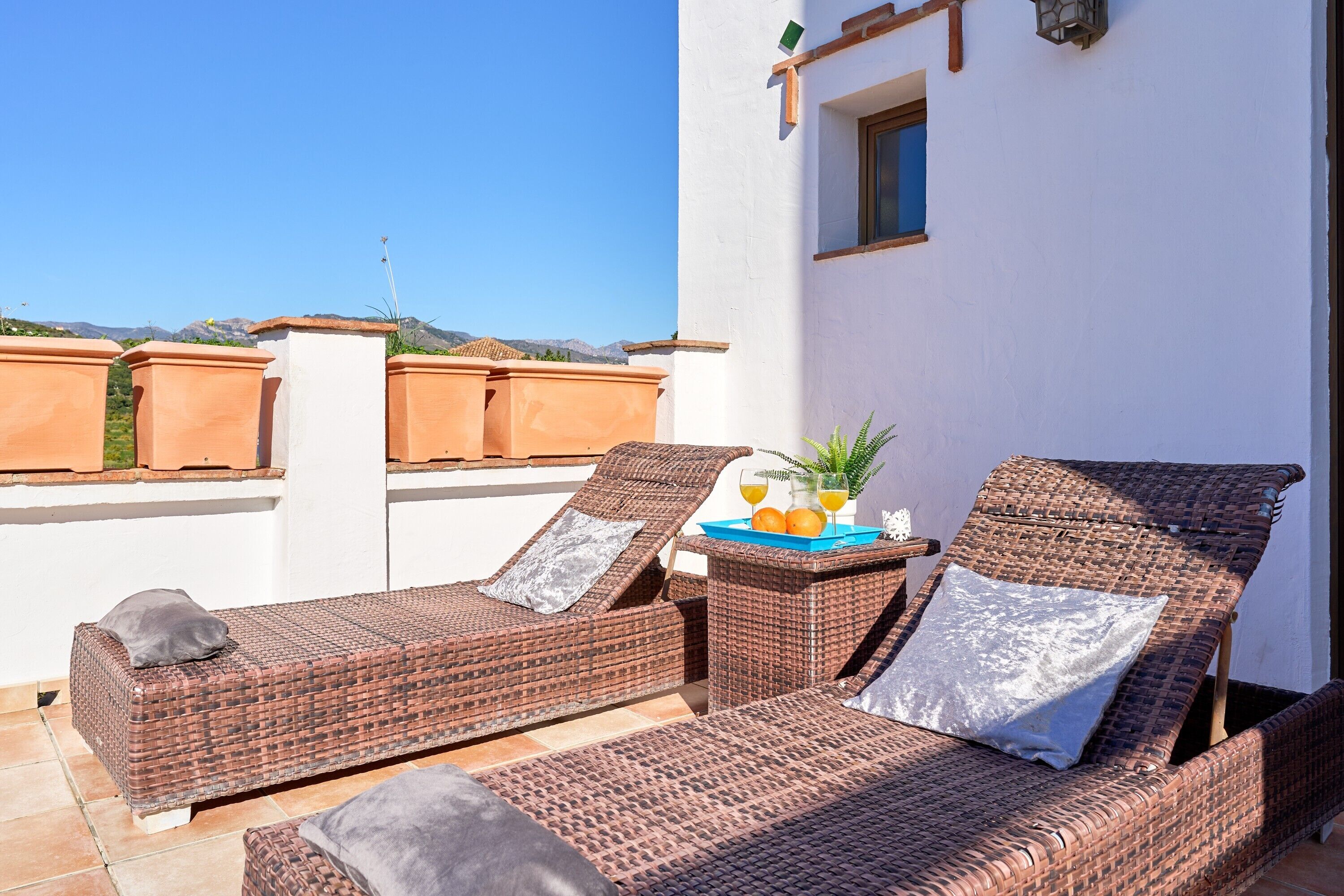Una terraza en la azotea con sillones de mimbre, una mesa con una bandeja de frutas y bebidas, y macetas de terracota.
