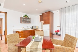 Un espace de vie compact comprenant un coin cuisine, un coin repas avec des meubles en osier et une petite table d’appoint agrémentée d’une plante en pot.