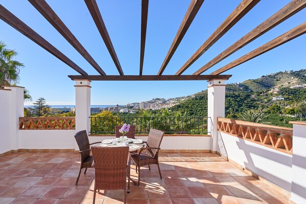 A covered outdoor patio with a table and chairs, a view of a hillside, and a railing.