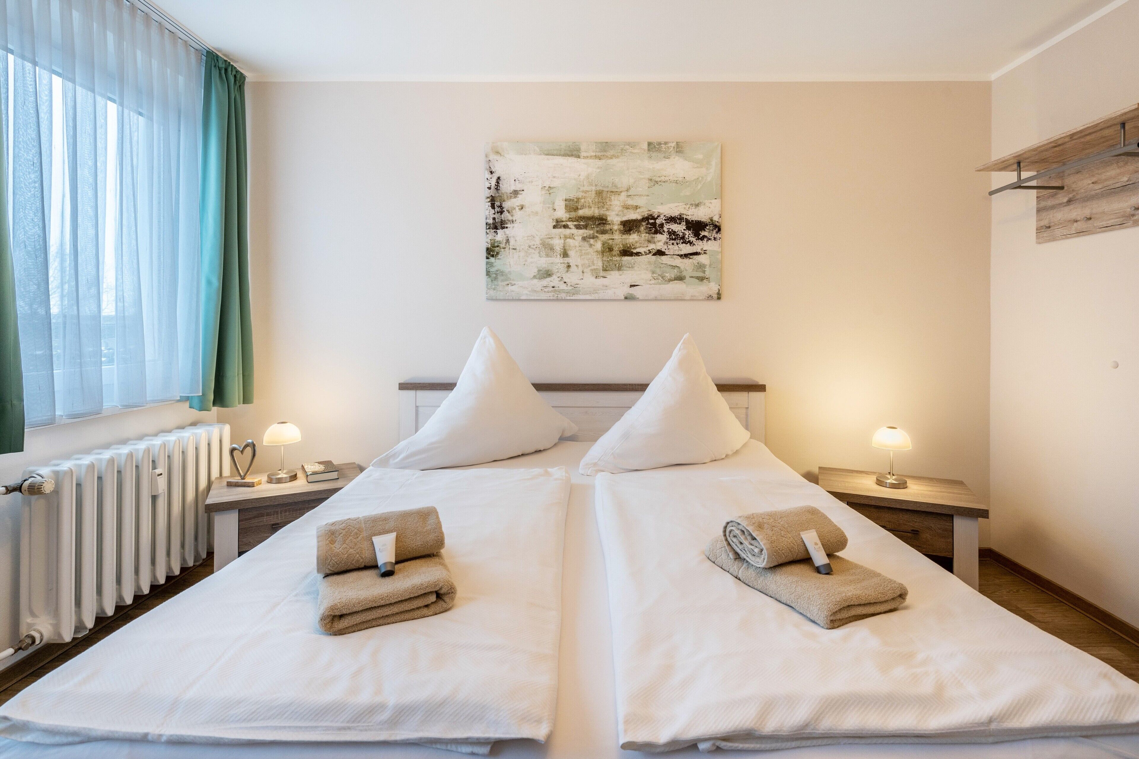 Ein Doppelzimmer mit zwei Betten, einem Heizkörper, einem Fenster mit Vorhängen, einem an der Wand hängenden abstrakten Gemälde und Nachttischlampen.