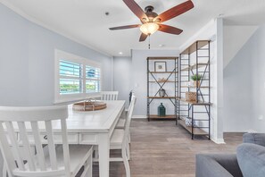 Een eetruimte met een witte tafel en stoelen, een plafondventilator en een wandplank met decoratieve objecten.