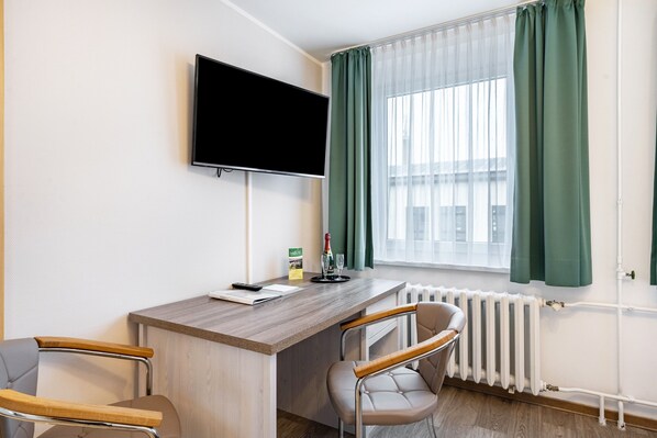Une chambre d’hôtel équipée d’un téléviseur à écran plat, d’un bureau en bois, de deux chaises, d’un radiateur et d’une fenêtre avec des rideaux verts.