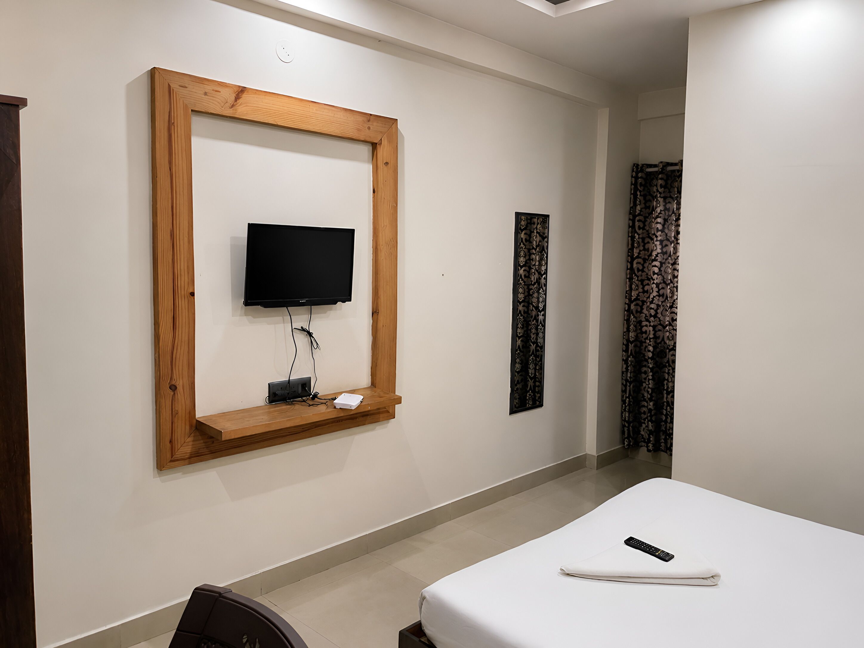 Habitación de hotel con televisor montado, estante de madera con toma de corriente, cama con control remoto y cortina estampada.