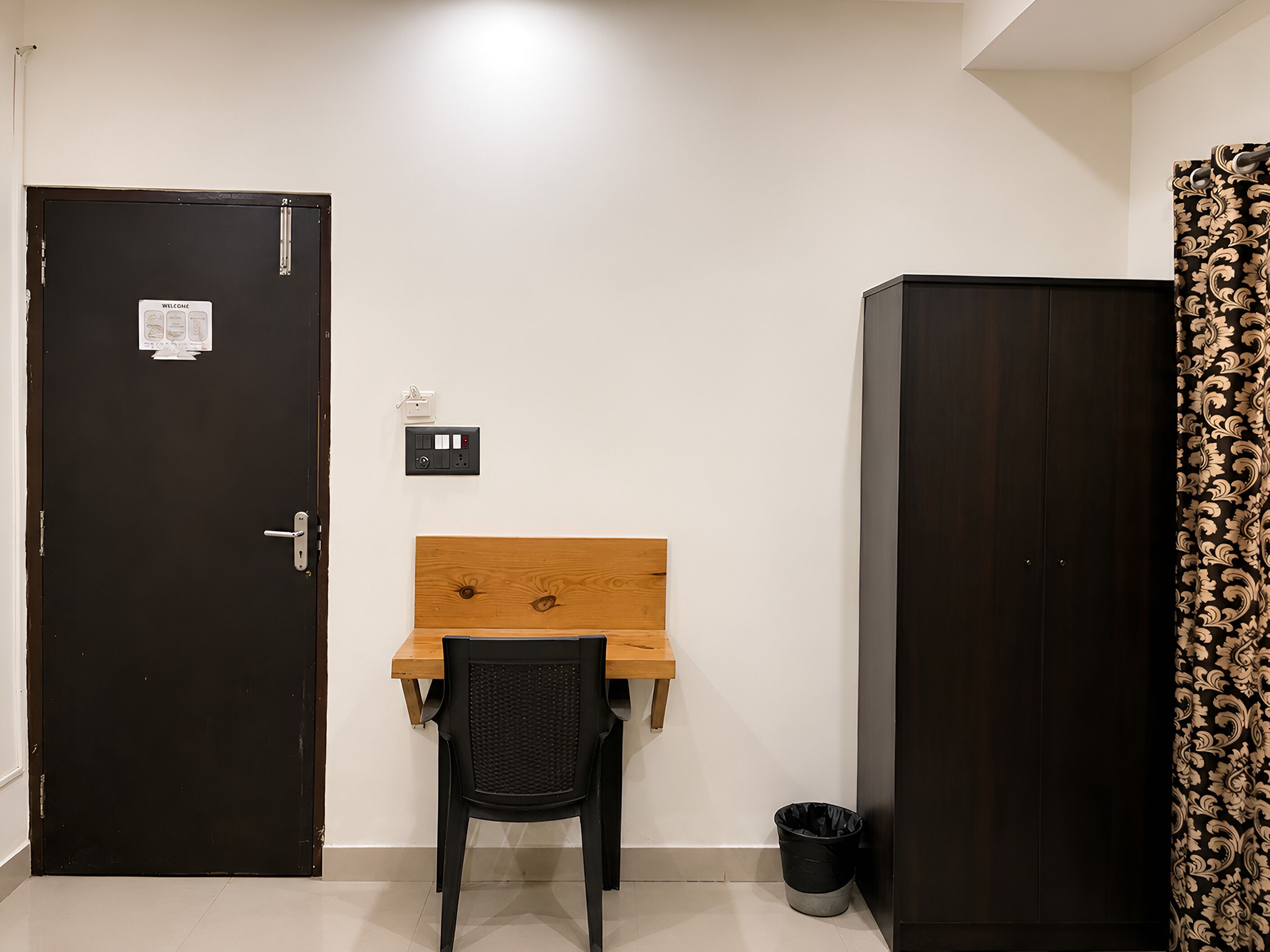 A room with a wooden desk, a black chair, a black trash bin, a door with a welcome sign, and a patterned curtain.