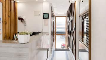 A reception area with a reception desk, a potted plant, a wall-mounted fan, and a view of the street through a window.