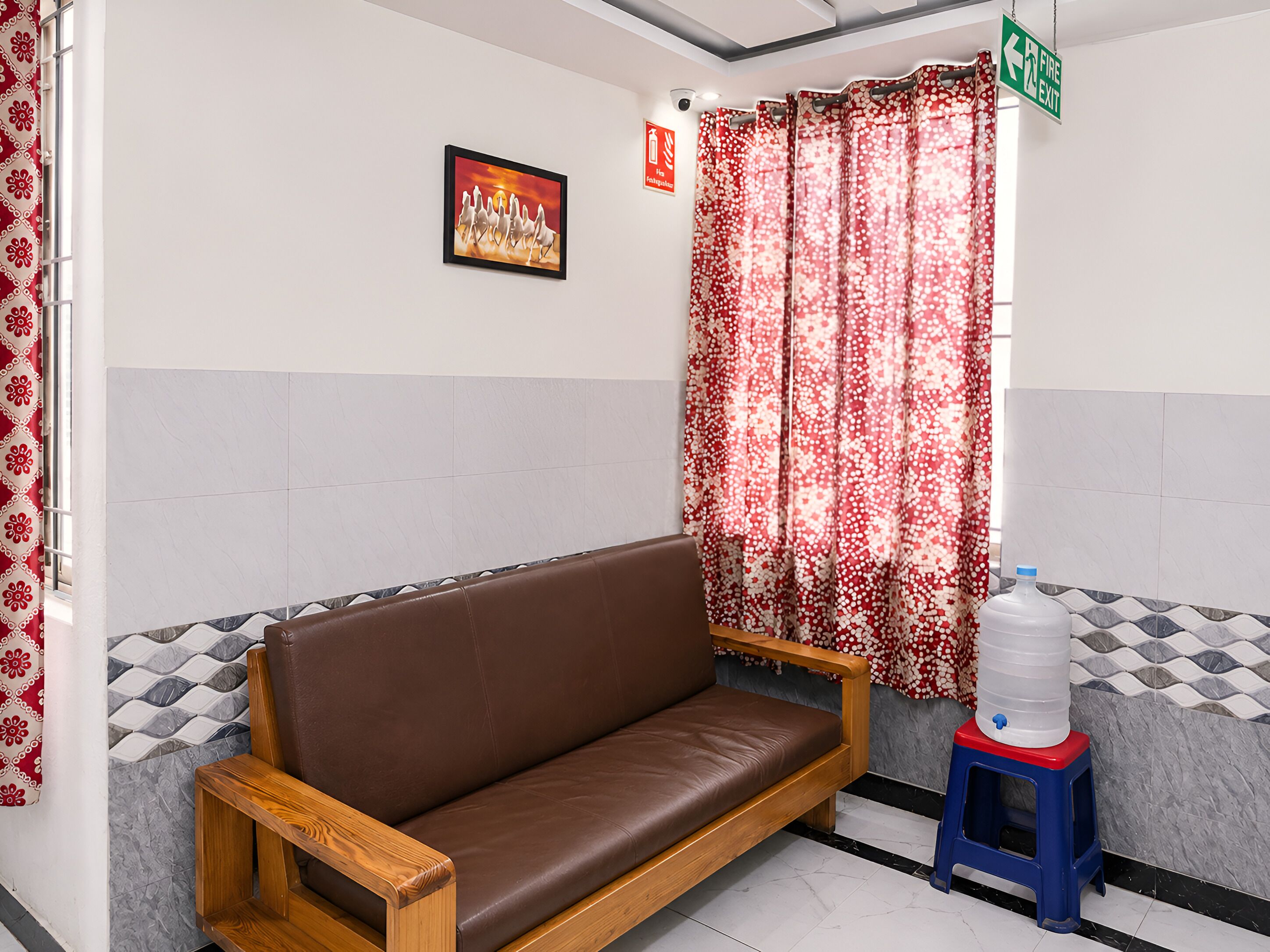 A brown leather sofa, a water dispenser, and a fire exit sign in a room with patterned tiles and a floral curtain.