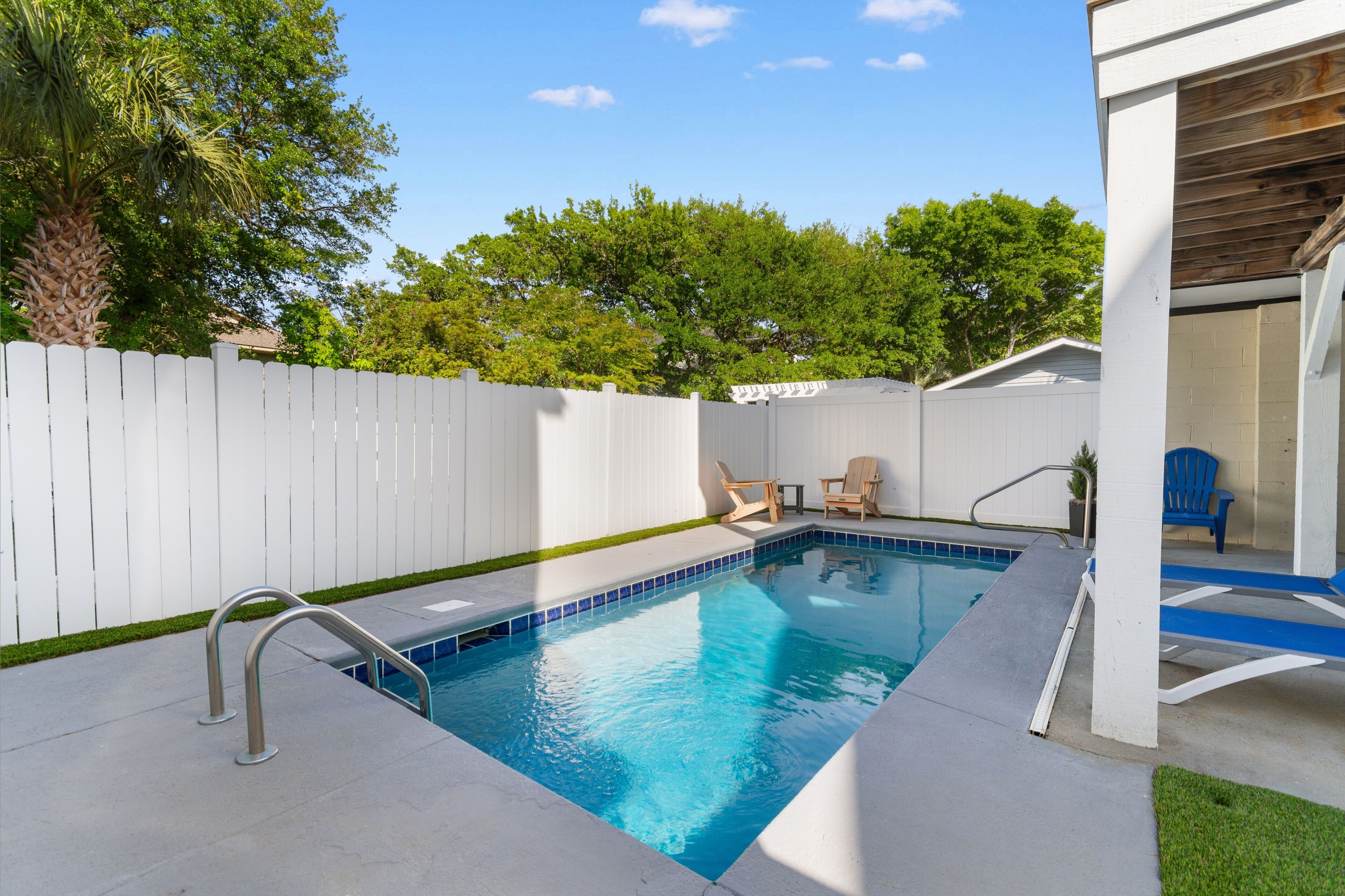 A swimming pool with a white fence, a covered area with chairs, and a view of trees.
