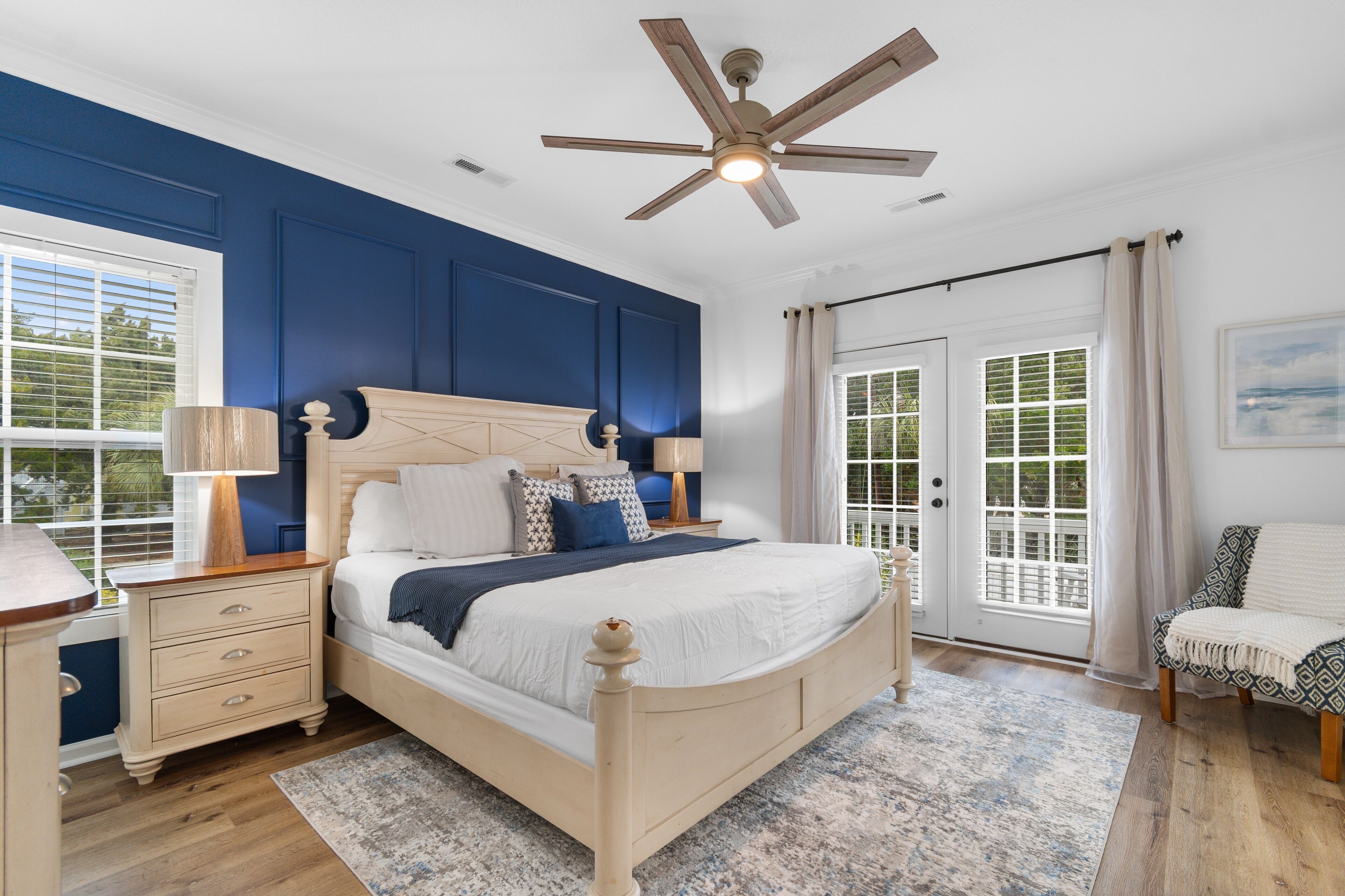 A bedroom with a large bed, a ceiling fan, a nightstand, a chair, and a view of the outdoors through a sliding door.