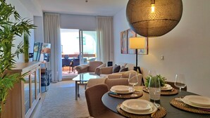 En moderne stue med sofa, spisebord, stole, et fjernsyn og et stort vindue med gardiner.