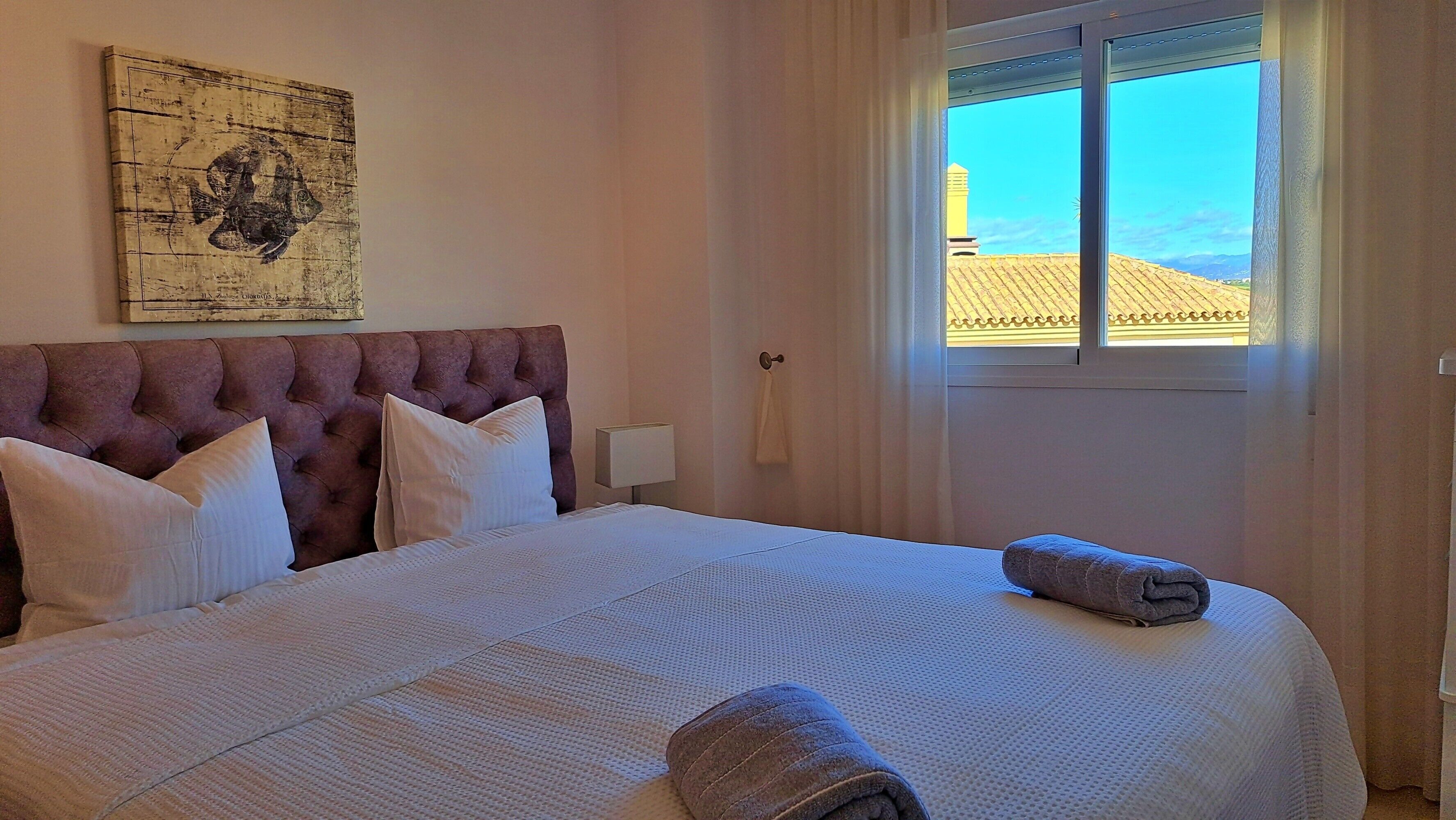 A neatly made bed with white linens, a tufted headboard, and a rolled towel on the side.