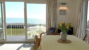 Uma sala de jantar com uma mesa posta para dois, um vaso com flores e vista para o mar através de uma porta de vidro.