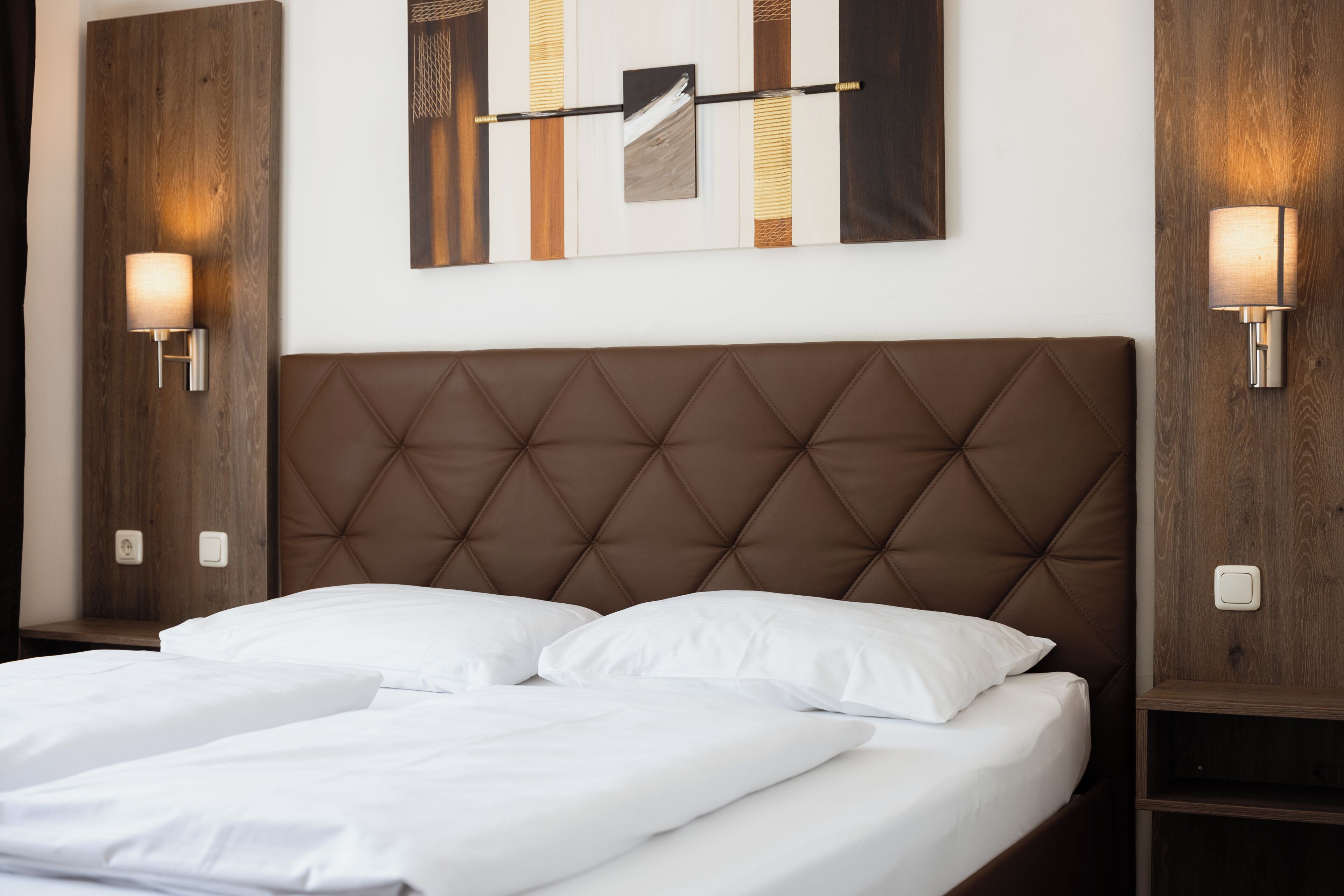 Quarto de hotel com cabeceira marrom acolchoada, duas camas brancas e luminárias fixadas na parede.