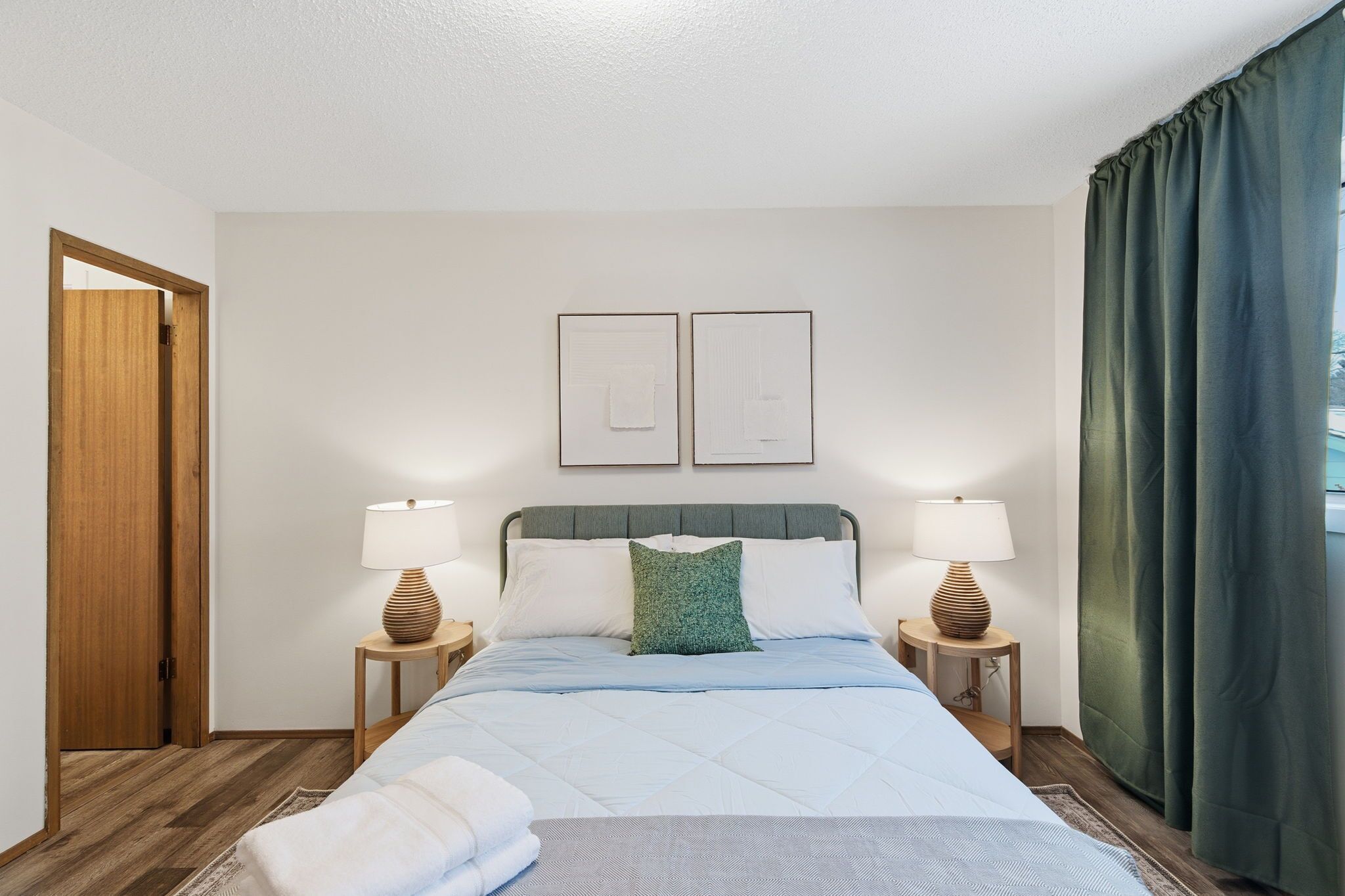 A neatly made bed with a blue bedspread, two bedside lamps, a green pillow, and two framed pictures on the wall.
