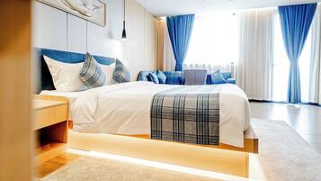 A modern hotel room with a bed, bedside tables, a desk, and a view of the outdoors through large windows.