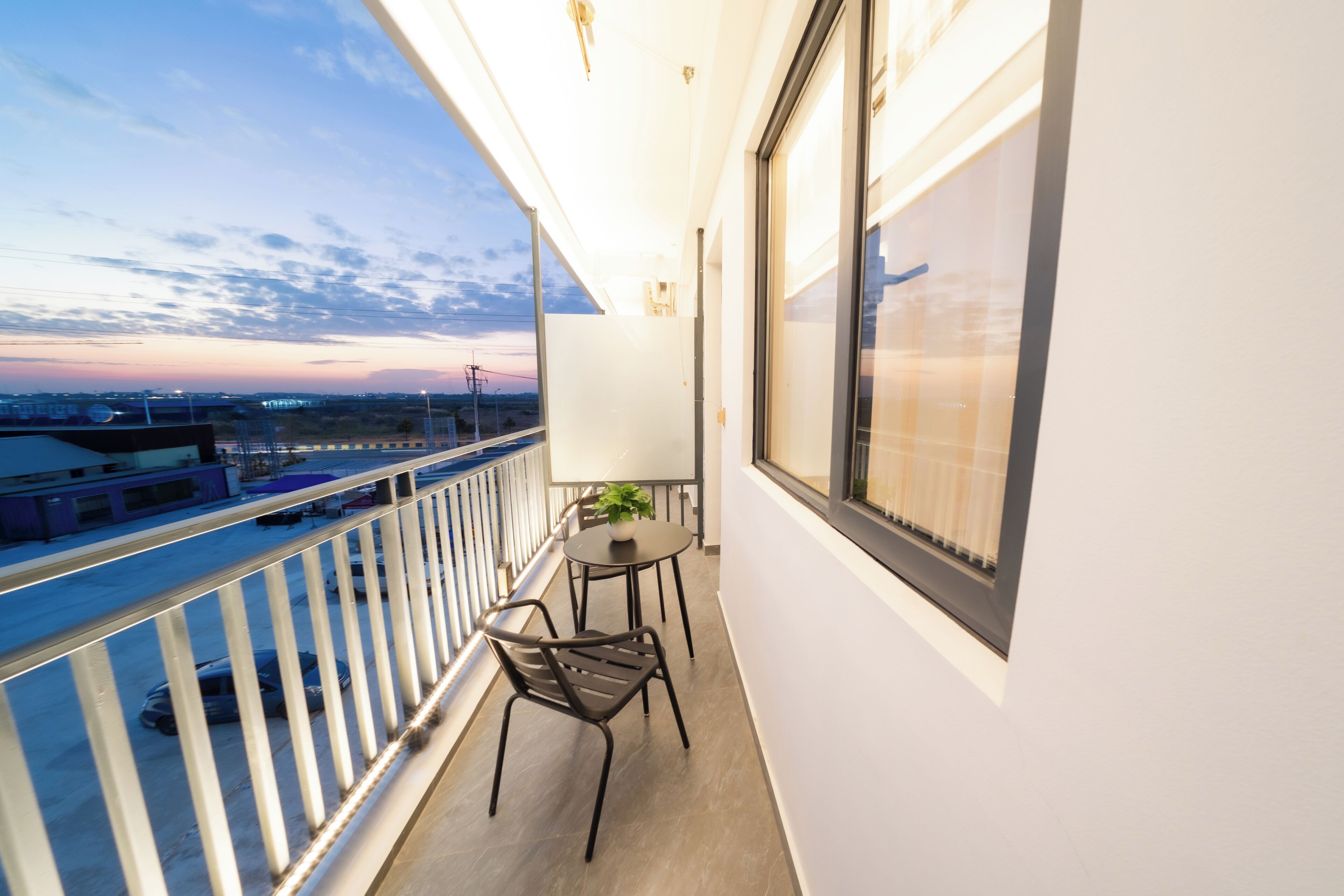 A balcony with a table and chairs, a potted plant, and a view of the cityscape at dusk.