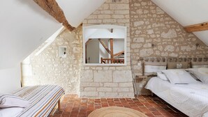 A bedroom with a sloped ceiling, exposed wooden beams, a stone wall, a bed with white bedding, and a mirror above the bed.
