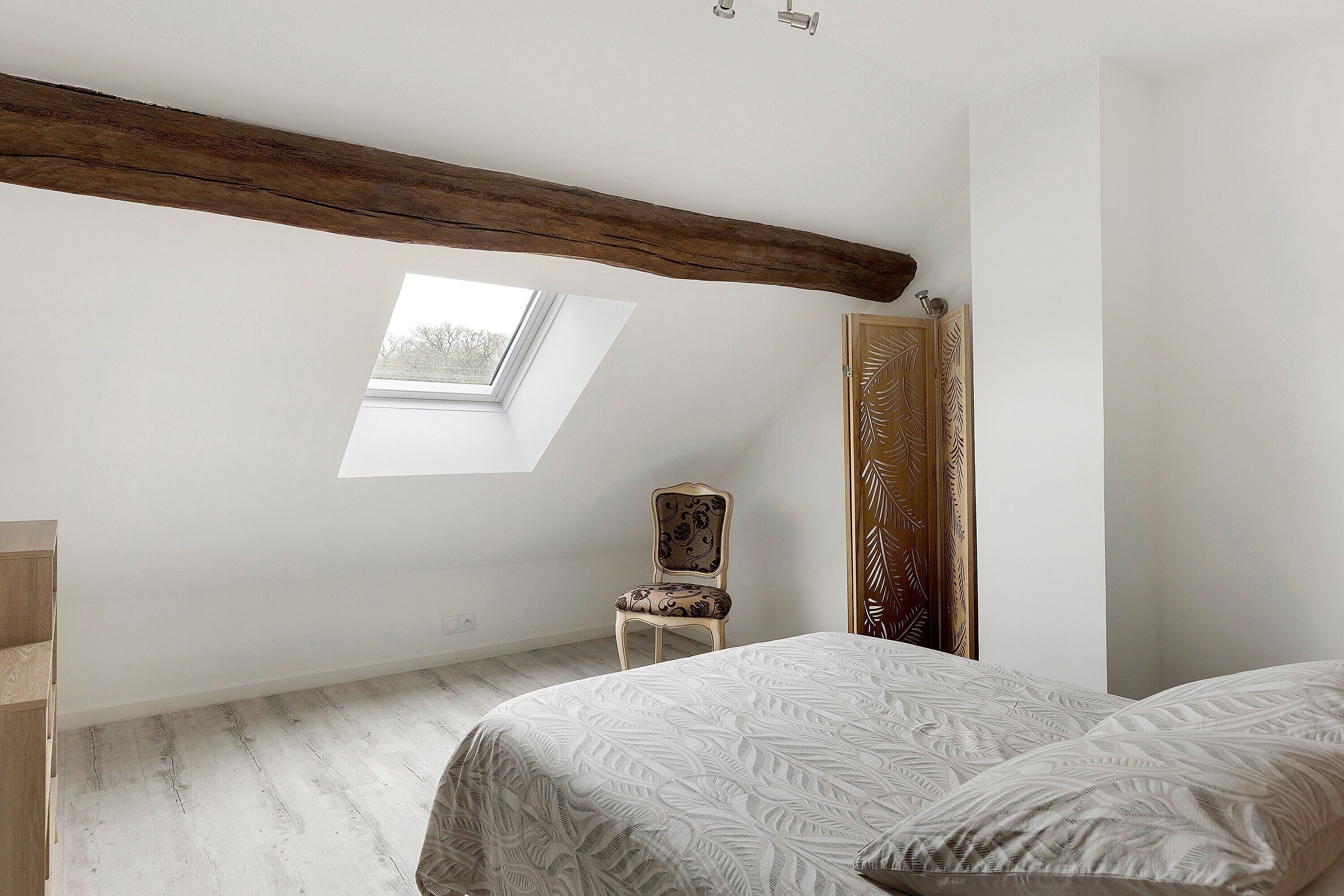 A bedroom with a sloped ceiling, a skylight, a wooden chair, a bed with patterned bedding, and a wardrobe with a leaf-patterned door.