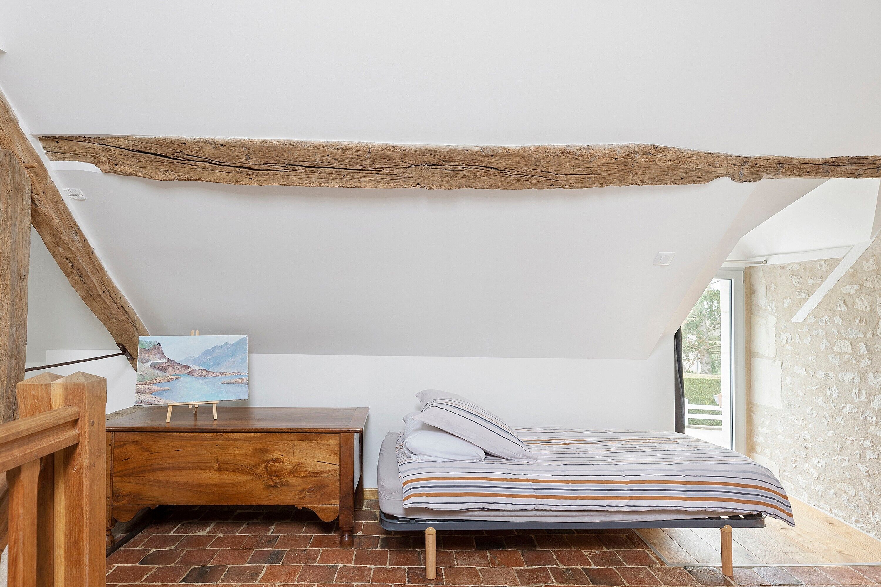 A bedroom with a sloped ceiling, a wooden desk, a bed with striped bedding, and a painting on the wall.