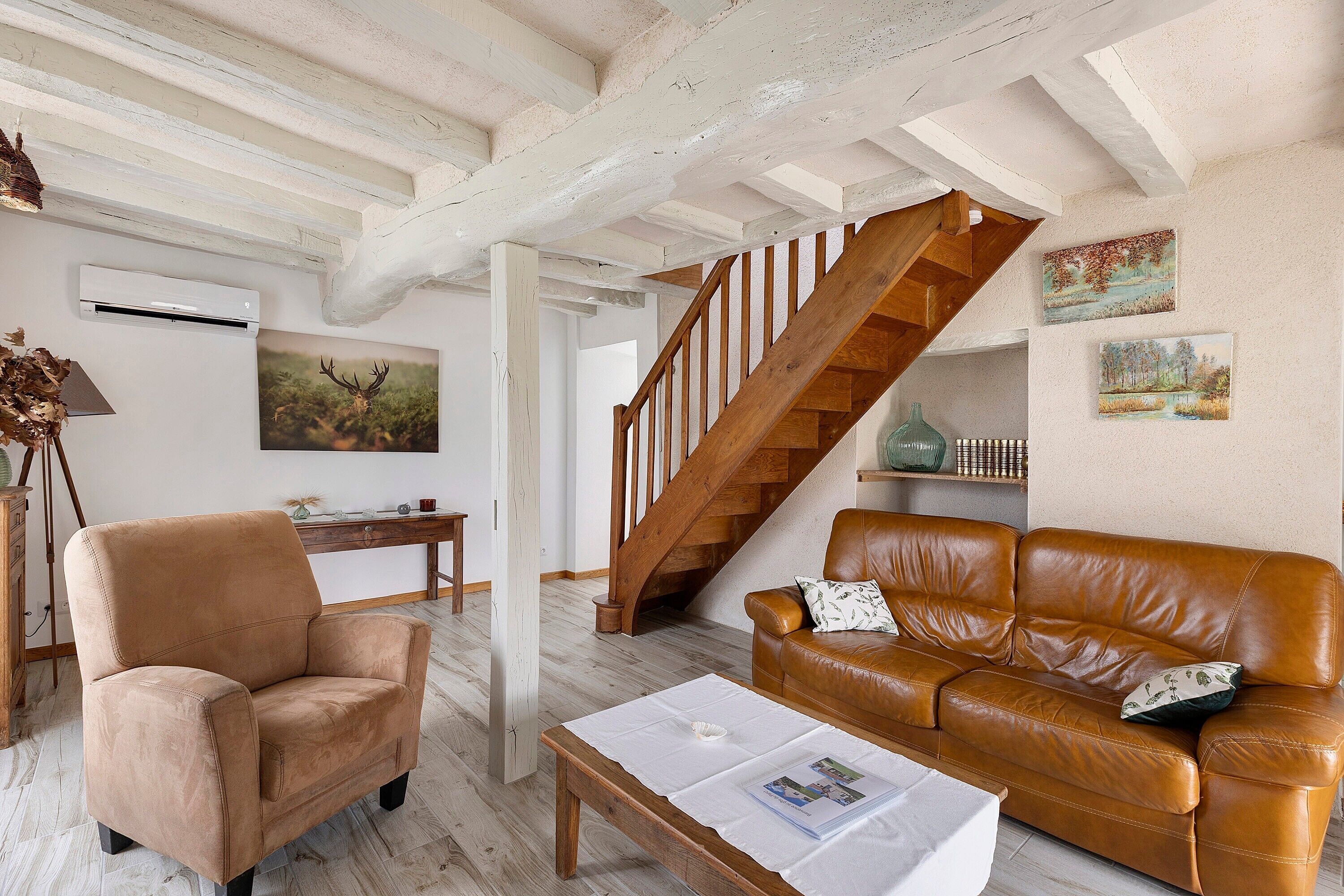 A living room with a brown leather sofa, a wooden coffee table, a brown armchair, a wooden staircase, and two framed paintings on the wall.
