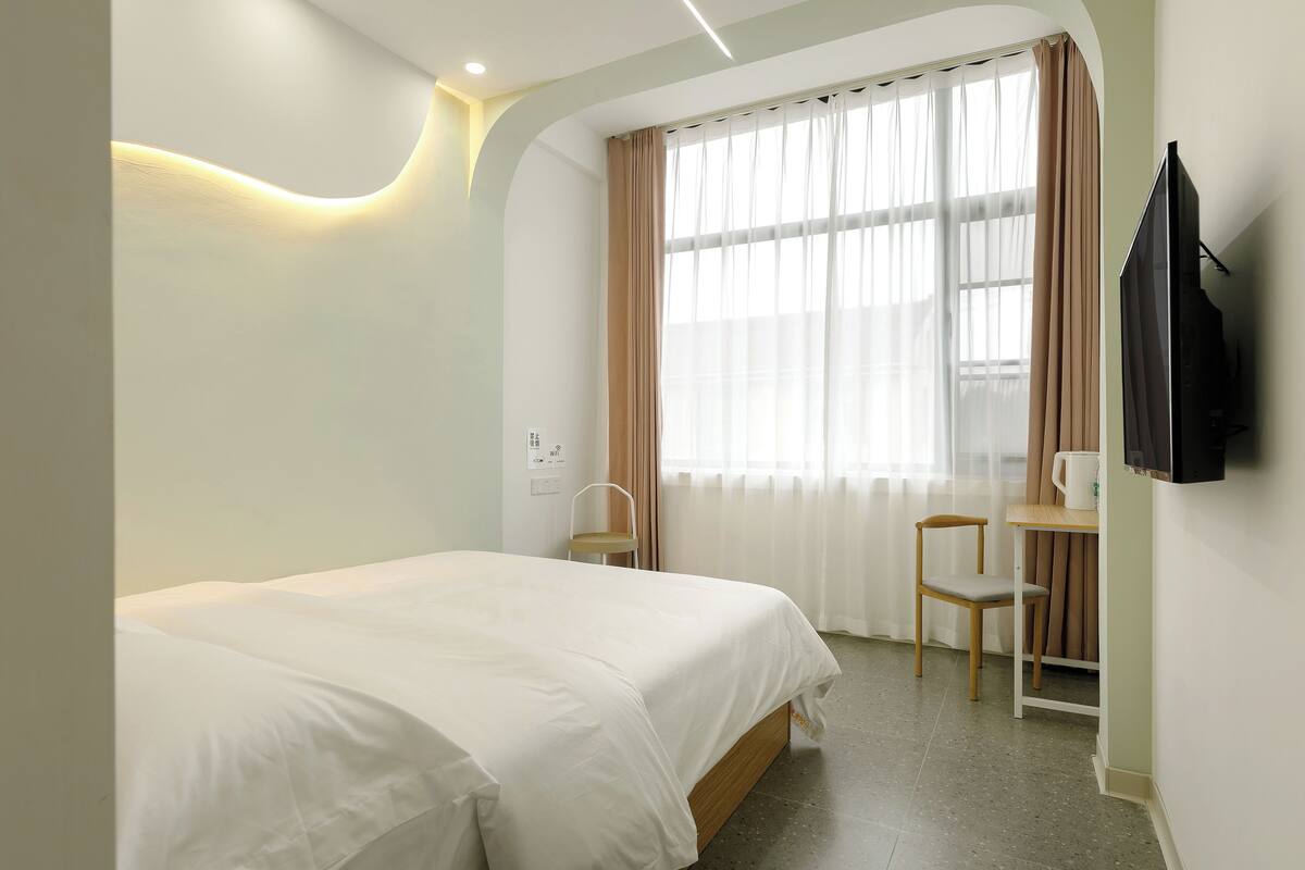 Habitación de hotel con cama, escritorio, silla, televisor y ventana con cortinas.