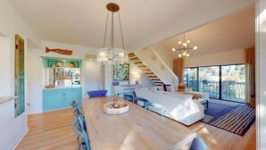 Un'ampia zona giorno con un tavolo da pranzo in legno, sedie blu, un divano bianco e una scala che conduce a un balcone.