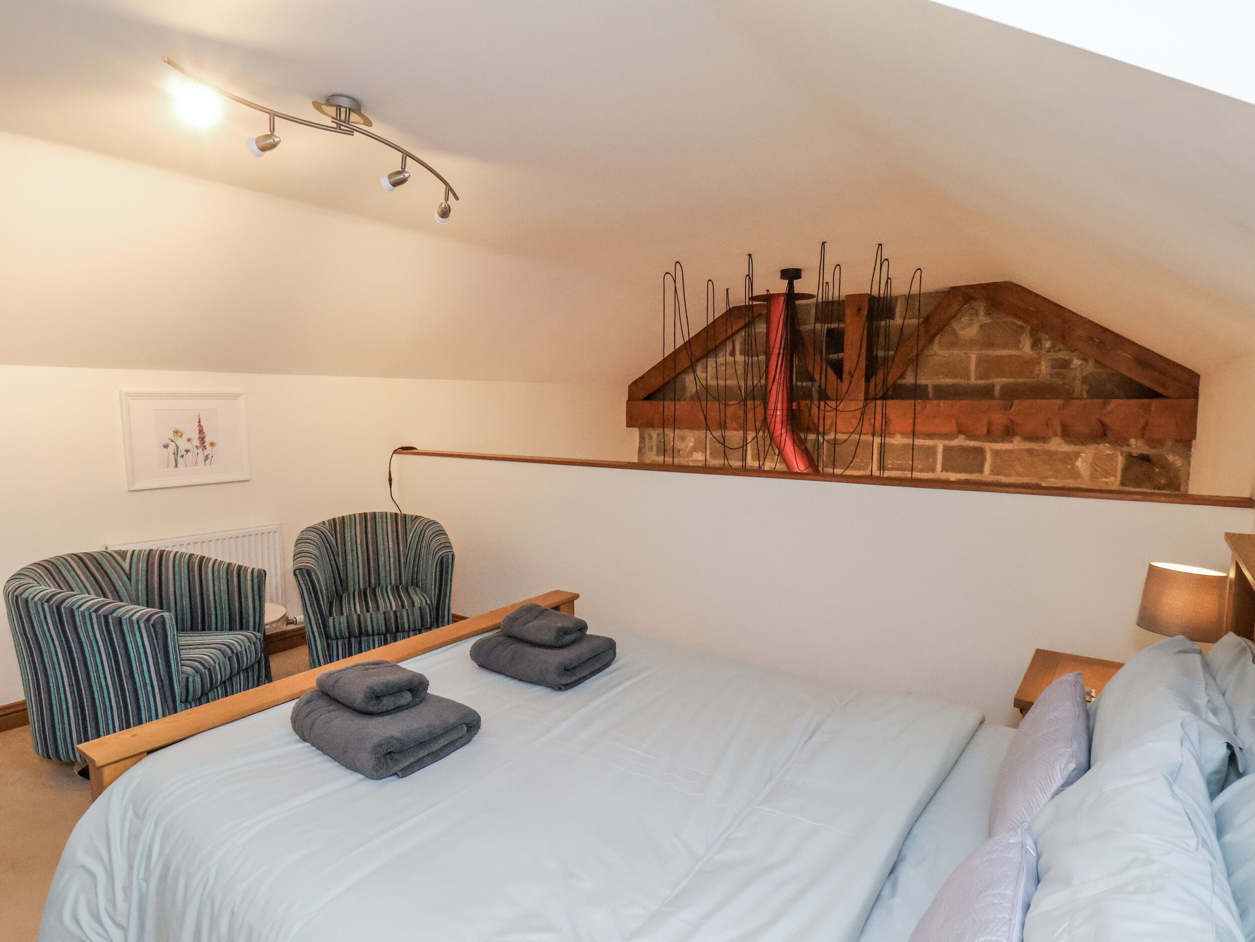 Um quarto com uma cama, duas poltronas listradas, um banco de madeira, um radiador, um quadro emoldurado e uma parede de tijolos com um cano vermelho.