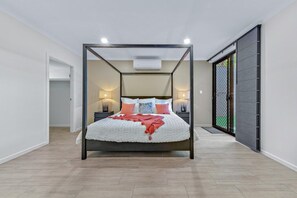 Ein Bett mit Baldachin, ausgestattet mit einer weißen und roten Tagesdecke, zwei Nachttischlampen und Blick auf eine grüne Außenanlage durch eine Schiebetür.