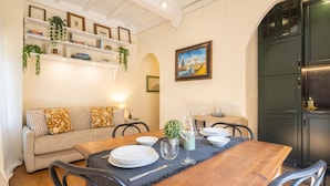 A dining area with a wooden table set for four, a sofa with patterned cushions, and shelves with books and plants. - Tornabuoni Cozy Apartment (Firenze)