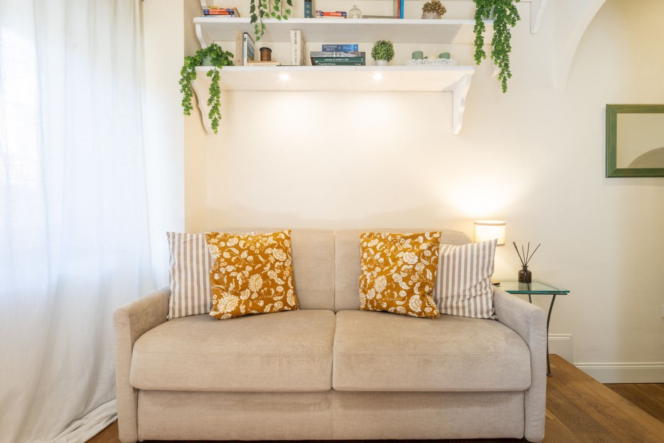 A beige sofa with floral cushions, a small side table with a lamp, and a bookshelf with books and plants.