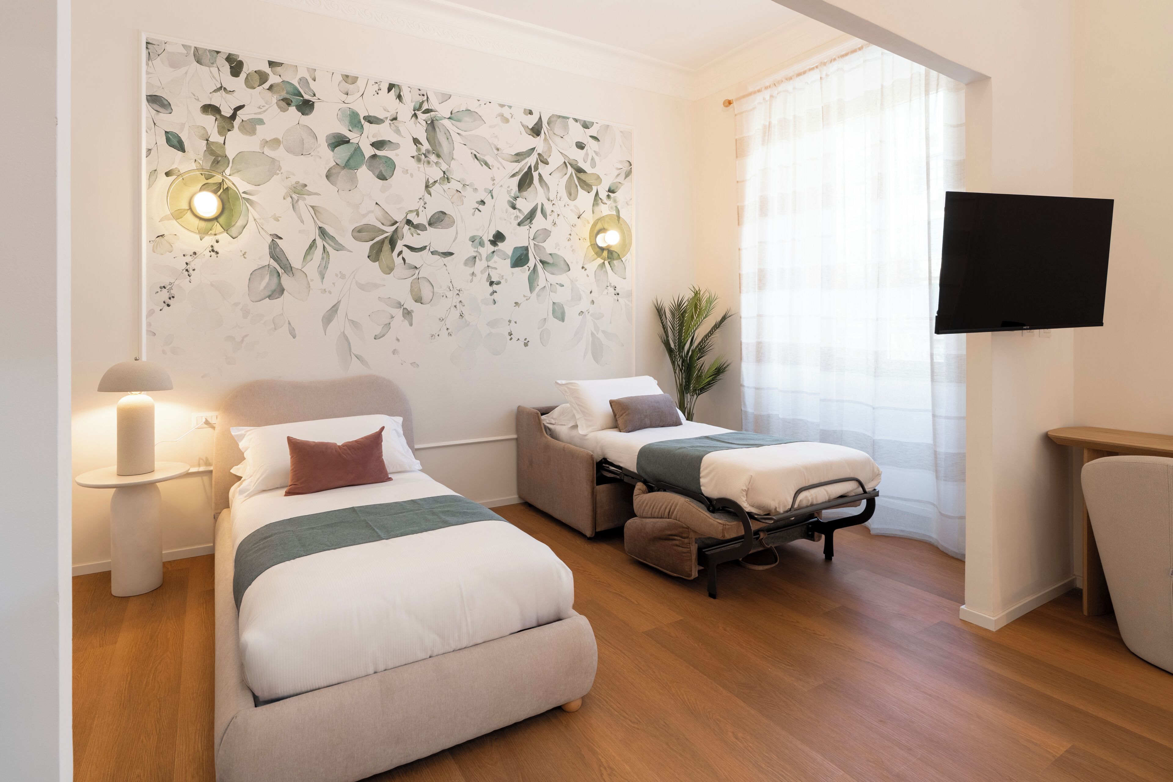 A hotel room with two beds, a flat-screen TV, a wall-mounted light fixture, and a potted plant.